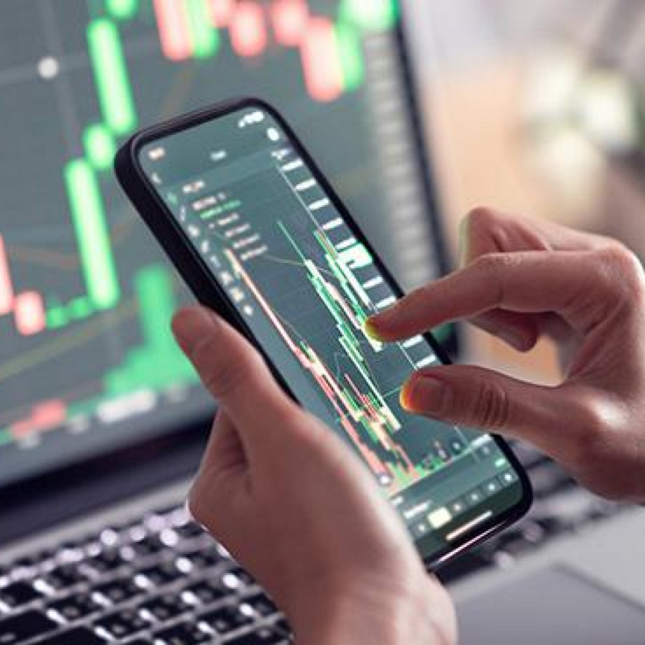 A hand zooming in on a graph displayed on a mobile phone screen and a laptop monitor showing another graph.