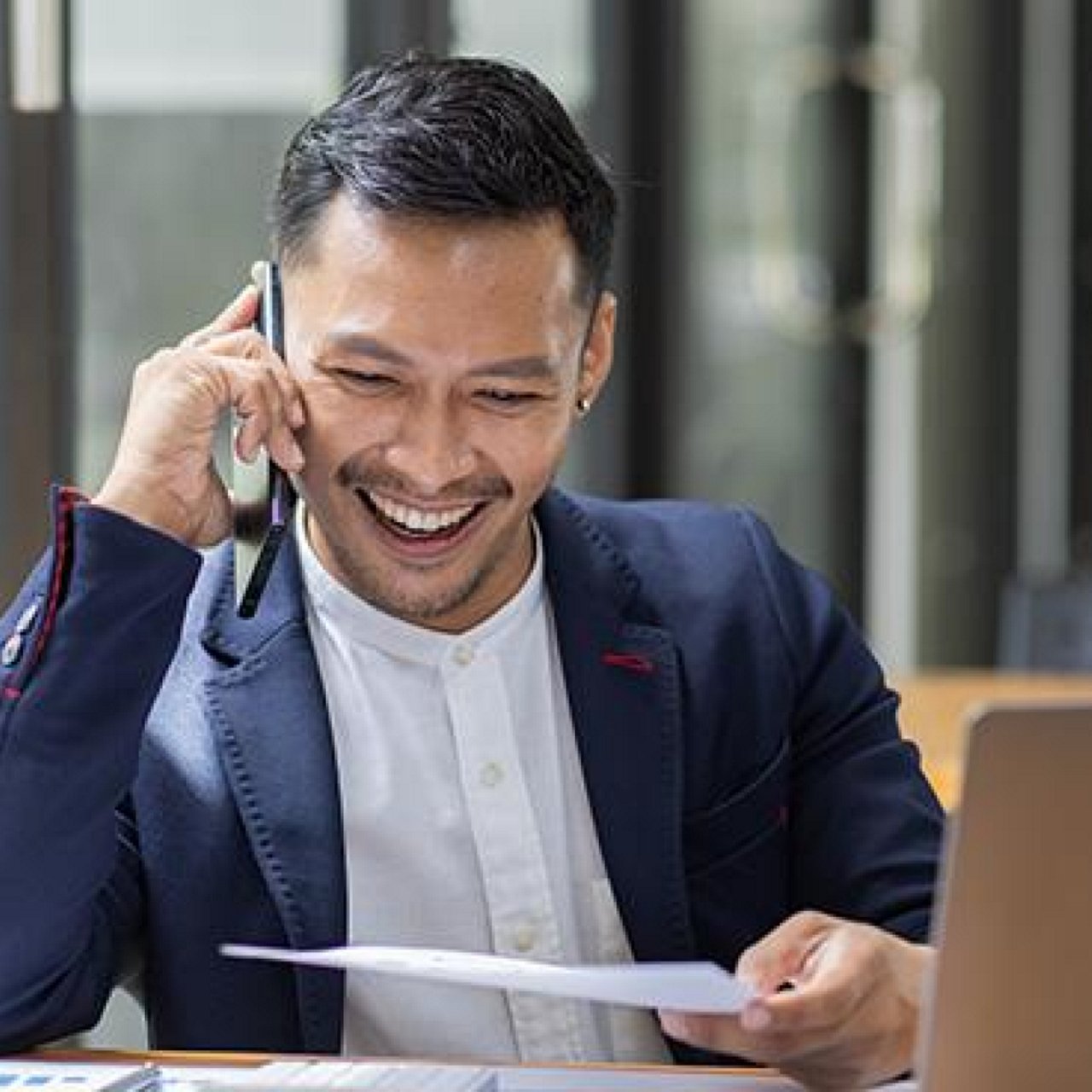 A man smiling while using his mobile phone with a laptop in front of him.