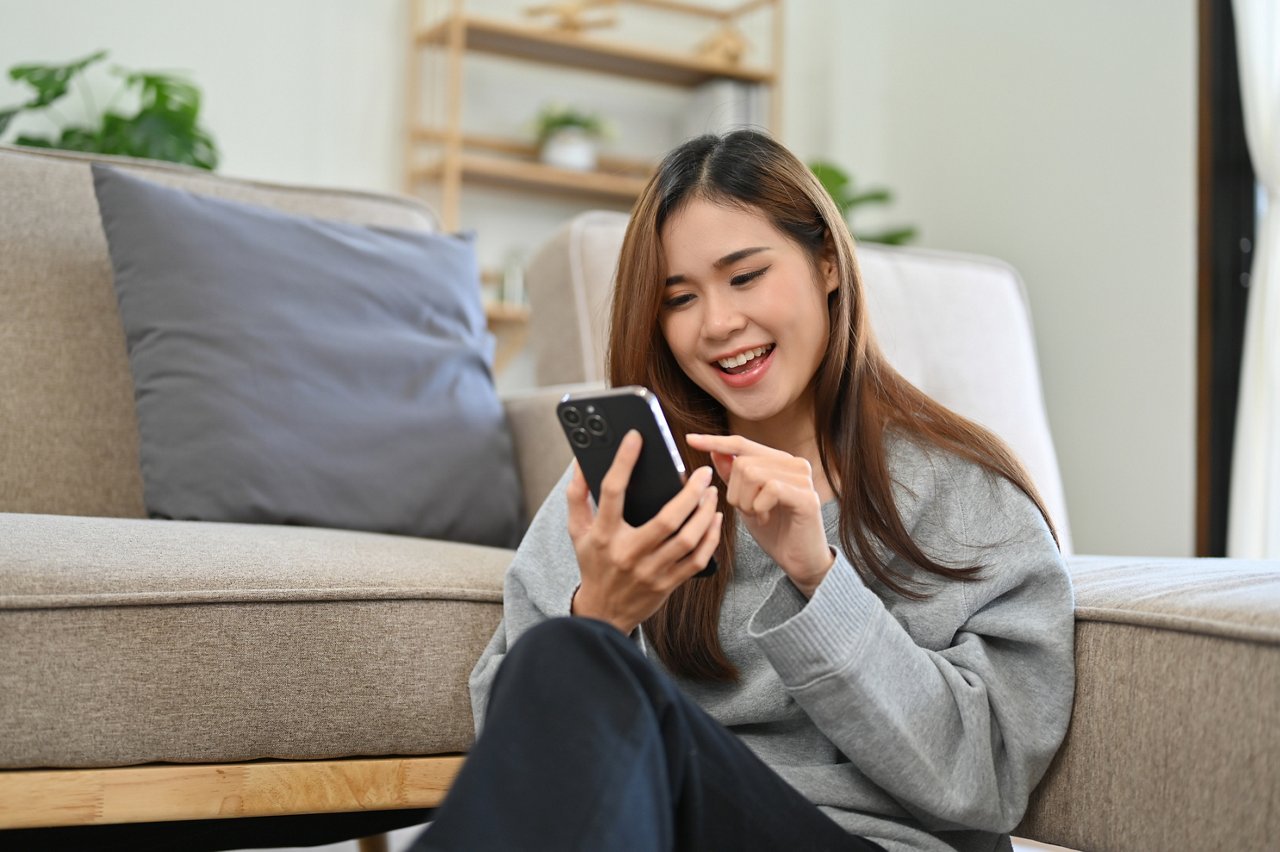 Attractive young Asian woman enjoys chatting with her friends on her smartphone while relaxing in her comfortable and minimal living room.
