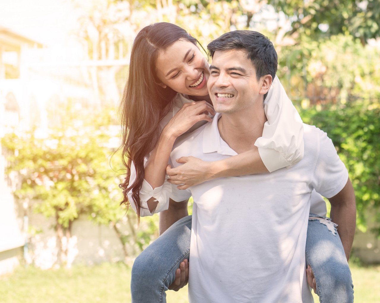 Portrait of cute romantic caucasian guy in checkered shirt, dreamy asian lady rides him on rear. Leisure, chill happiness, lawn stroll, relax, romance lifestyle, well dressed partners posing
