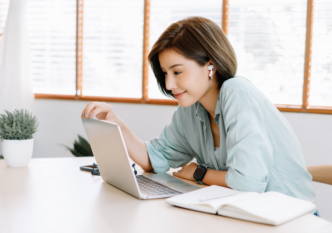 Happy young beautiful Asian woman using laptop working remote and wireless earphones sitting at table in home office.Remote work,E-learning, New normal concept.