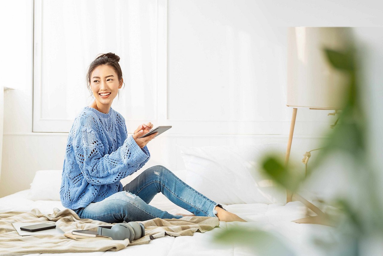 Smiling Asian woman scrolling through her tablet as she looks at a corner in a bright room.