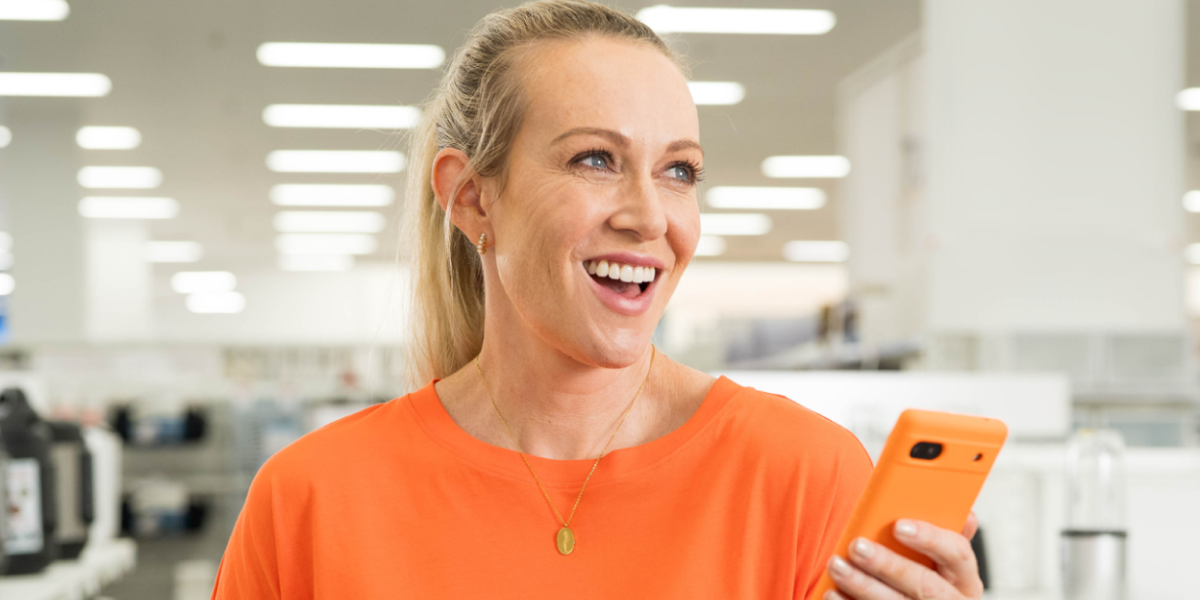 A smiling woman with blonde hair wearing an orange shirt holding an orange phone in a store setting.