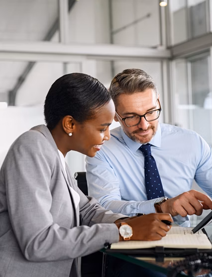 Mature businessman discussing work with black manager writing notes in diary. Confident business man in formal clothing working with african american woman showing data on digital tablet. Smiling professional business people working together on new business strategy.
