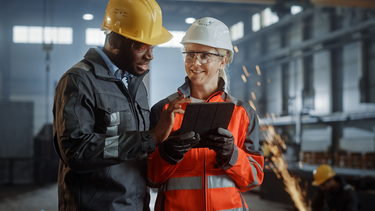 Two Heavy Industry Engineers Stand in Steel Metal Manufacturing Factory, Use Digital Tablet Computer and Have a Discussion. Black African American Industrial Specialist Talk to Female Technician.