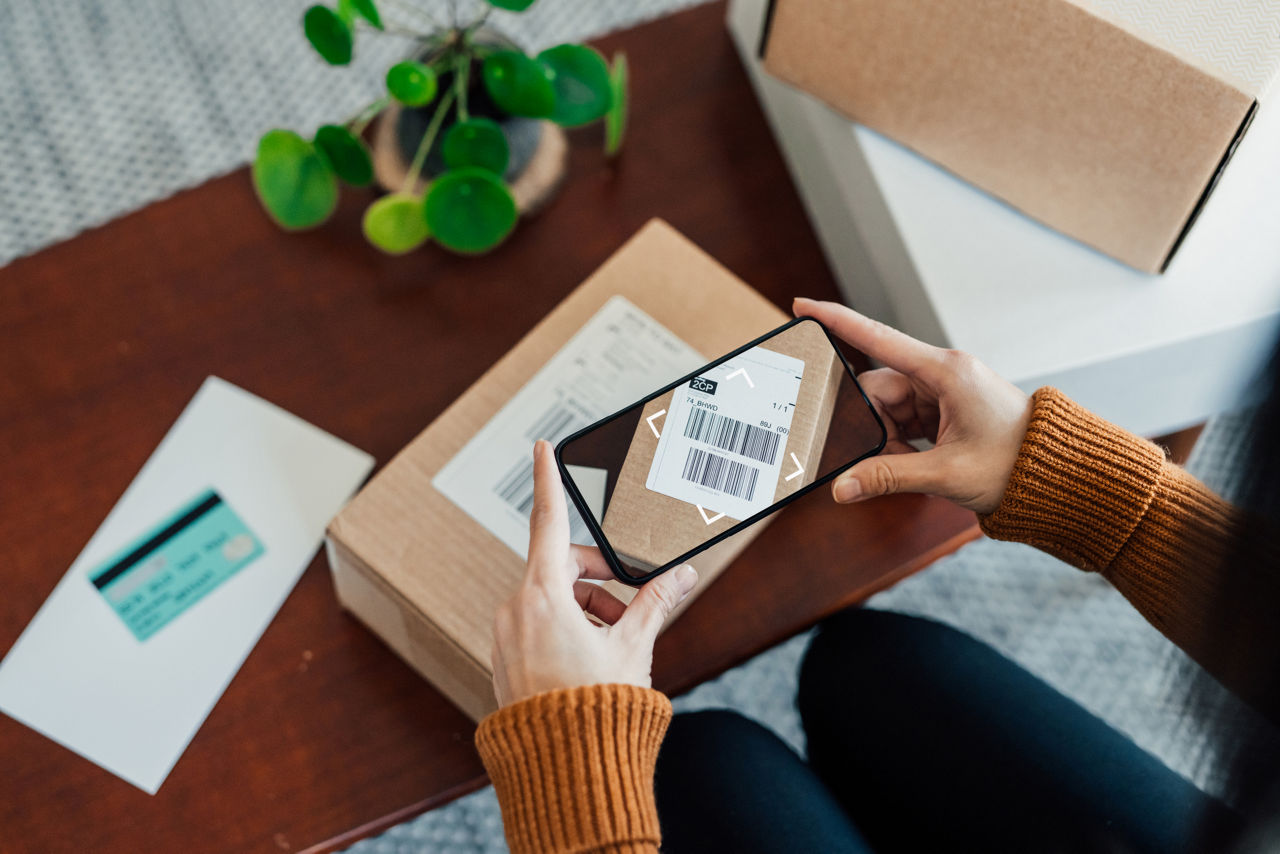 High angle view of small business owner working at home, scanning shipping label with smart phone. Shipping packages for e-commerce. Online shopping concept. Digital delivery concept.