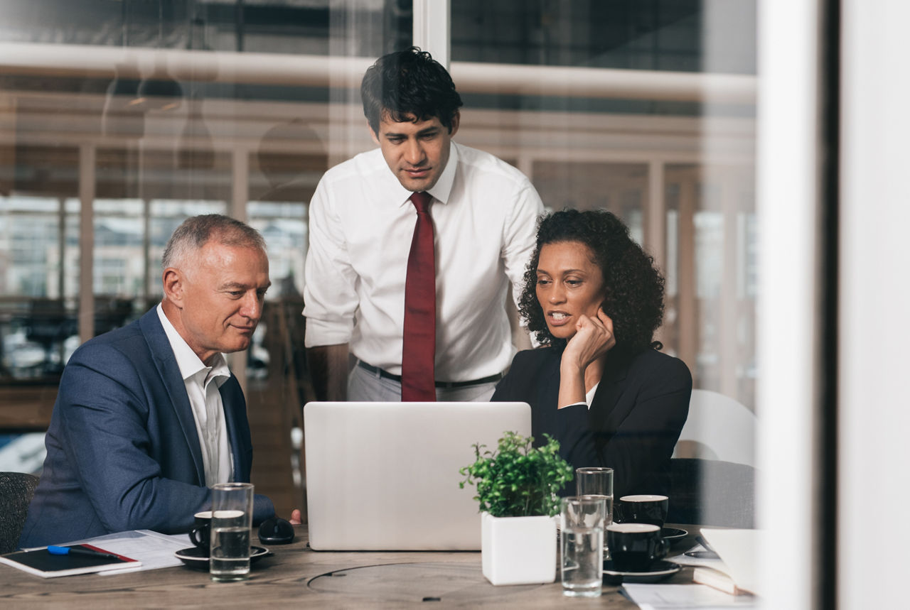 Three diverse businesspeople talking together over a laptop while working at a table in an office boardroom