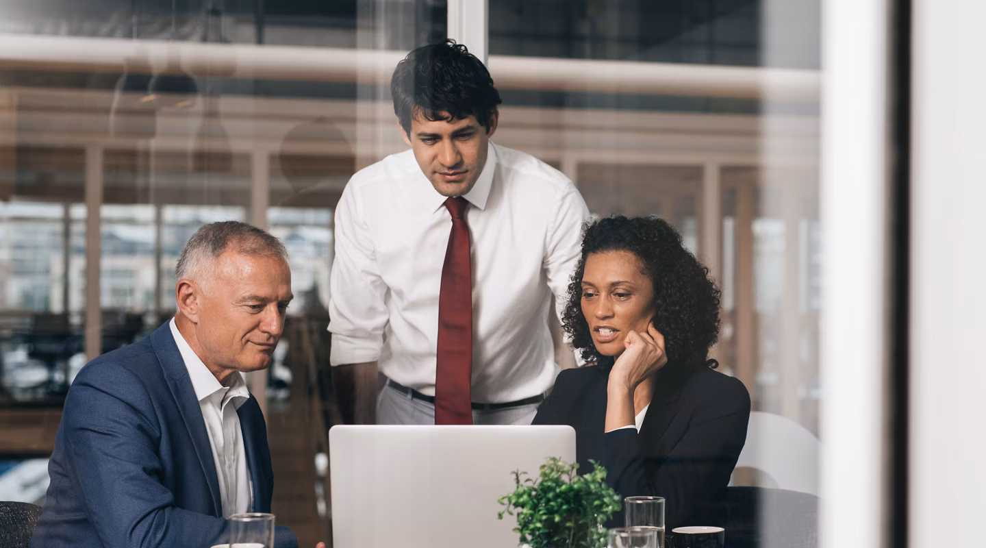 Three diverse businesspeople talking together over a laptop while working at a table in an office boardroom