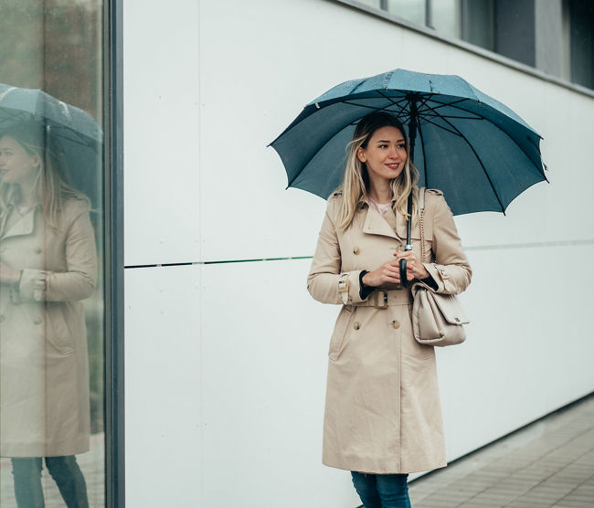 Young smiling businesswoman with umbrella and shoulder bag walking down city street during rain