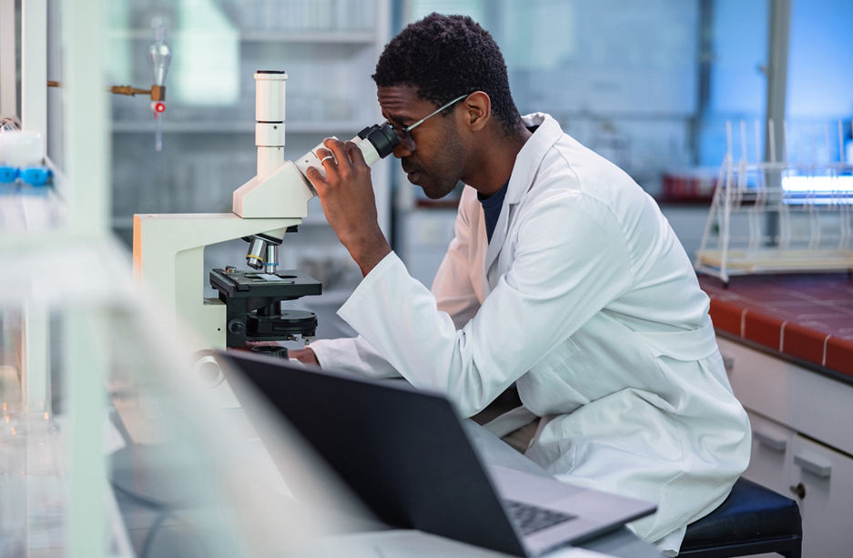 Portrait of African-American male scientist wearing a lab coat, looking under a microscope, analyzing a sample during his work in a laboratory.