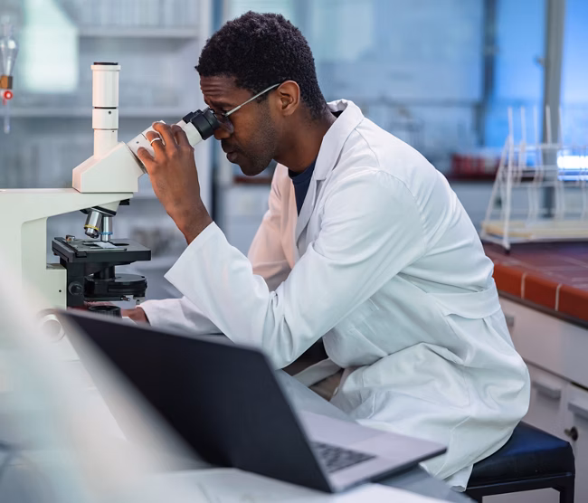 Portrait of African-American male scientist wearing a lab coat, looking under a microscope, analyzing a sample during his work in a laboratory.