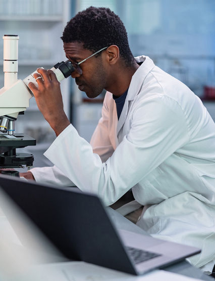 Portrait of African-American male scientist wearing a lab coat, looking under a microscope, analyzing a sample during his work in a laboratory.