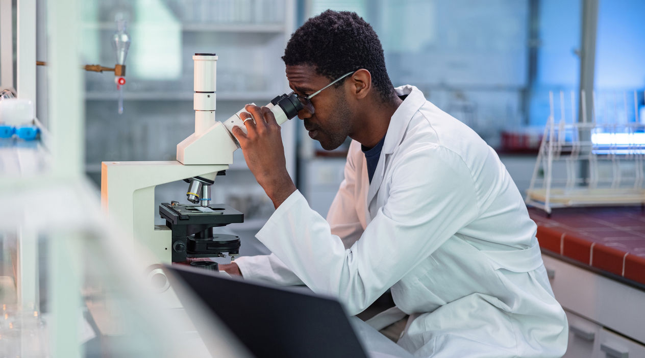 Portrait of African-American male scientist wearing a lab coat, looking under a microscope, analyzing a sample during his work in a laboratory.