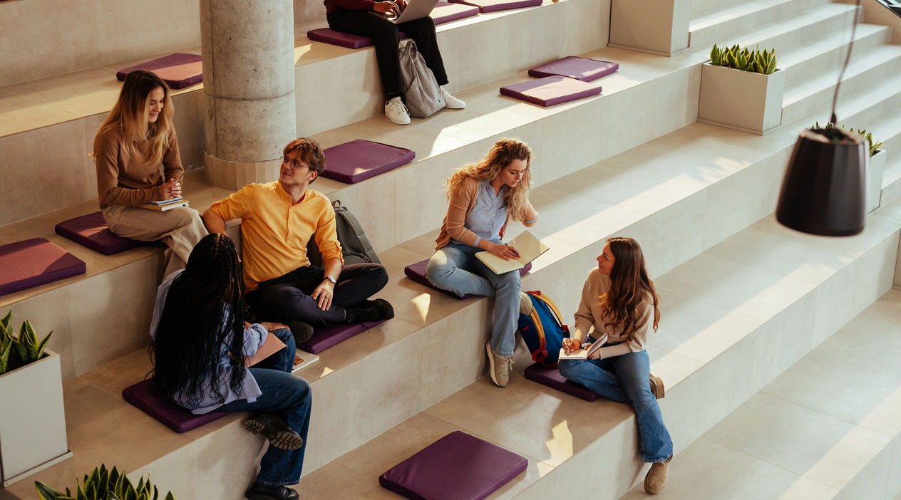 Diverse university students are studying and chatting, sitting on modern bleachers in the hall of their college