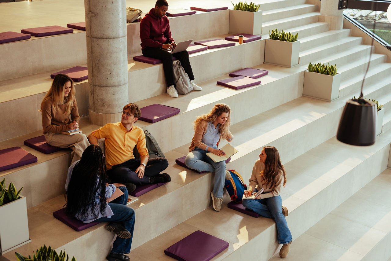Diverse university students are studying and chatting, sitting on modern bleachers in the hall of their college