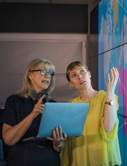 An experienced woman mentors a female colleague, the mature woman is holding a laptop as they debate data from an interactive display; they are both wearing smart casual clothing.