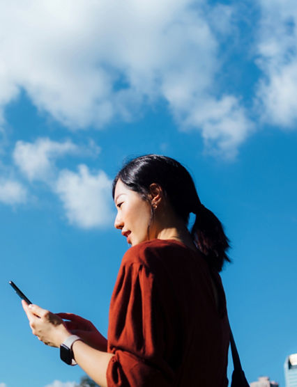 Low angle portrait of young Asian woman using smartphone against beautiful blue sky with cloudscapes, enjoying sunlight outdoors. Lifestyle and technology