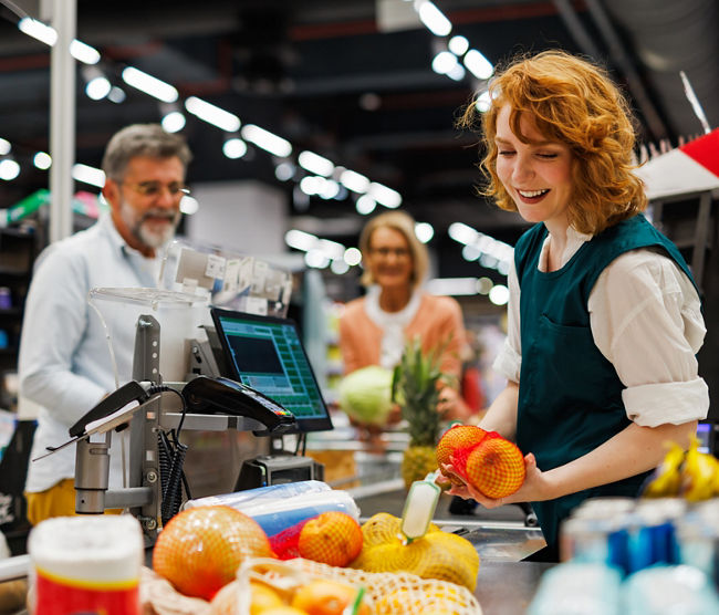 Smiling cashier scanning fruit at the checkout of a supermarket with customers waiting in the background, providing excellent customer service and efficient processing