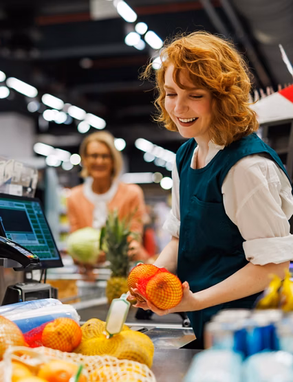 Smiling cashier scanning fruit at the checkout of a supermarket with customers waiting in the background, providing excellent customer service and efficient processing