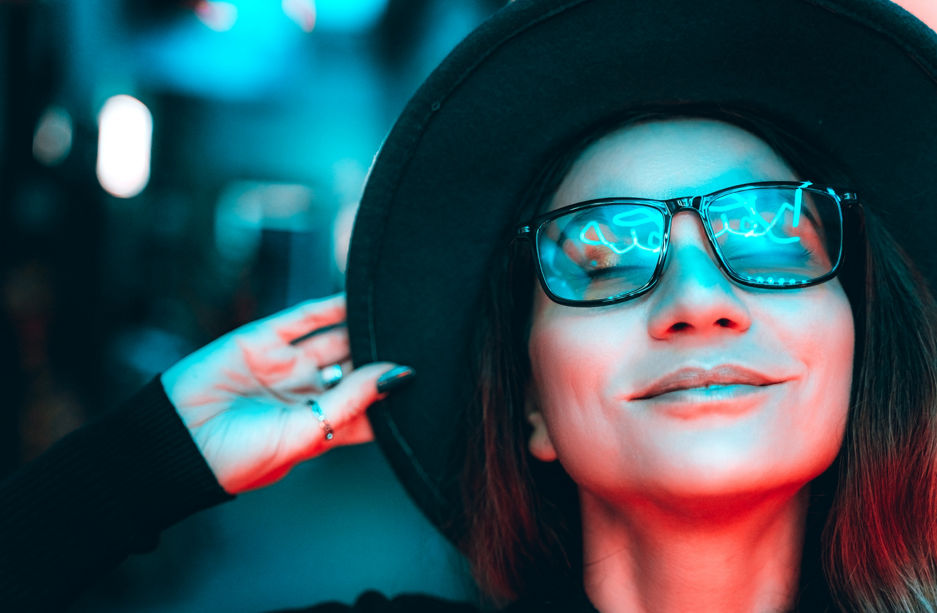 Portrait of a beautiful hipster woman in glasses and hat, she is dancing and dreaming with her eyes closed, enjoying the party night in illuminated neon lights of blue and red. The woman is smiling, reflections in her glasses. Space for text copy