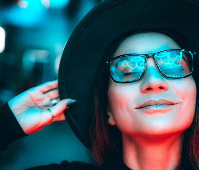 Portrait of a beautiful hipster woman in glasses and hat, she is dancing and dreaming with her eyes closed, enjoying the party night in illuminated neon lights of blue and red. The woman is smiling, reflections in her glasses. Space for text copy