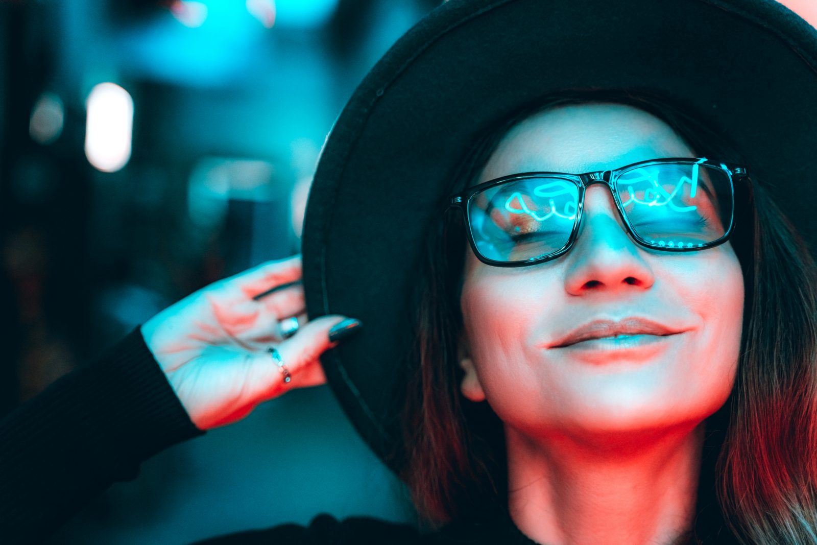 Portrait of a beautiful hipster woman in glasses and hat, she is dancing and dreaming with her eyes closed, enjoying the party night in illuminated neon lights of blue and red. The woman is smiling, reflections in her glasses. Space for text copy
