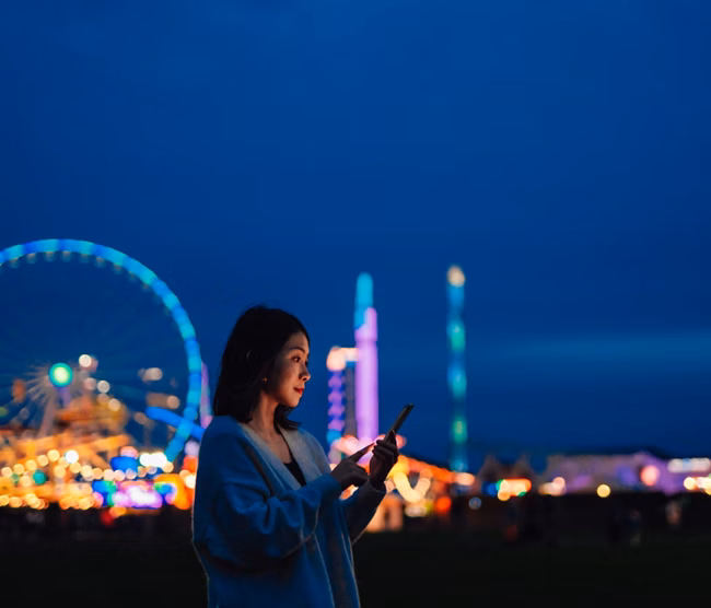 Side view of beautiful young Asian woman looking aways while using smartphone in carnival at night with illuminated night in the background. Mobile banking. Business on the go.