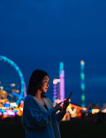 Side view of beautiful young Asian woman looking aways while using smartphone in carnival at night with illuminated night in the background. Mobile banking. Business on the go.