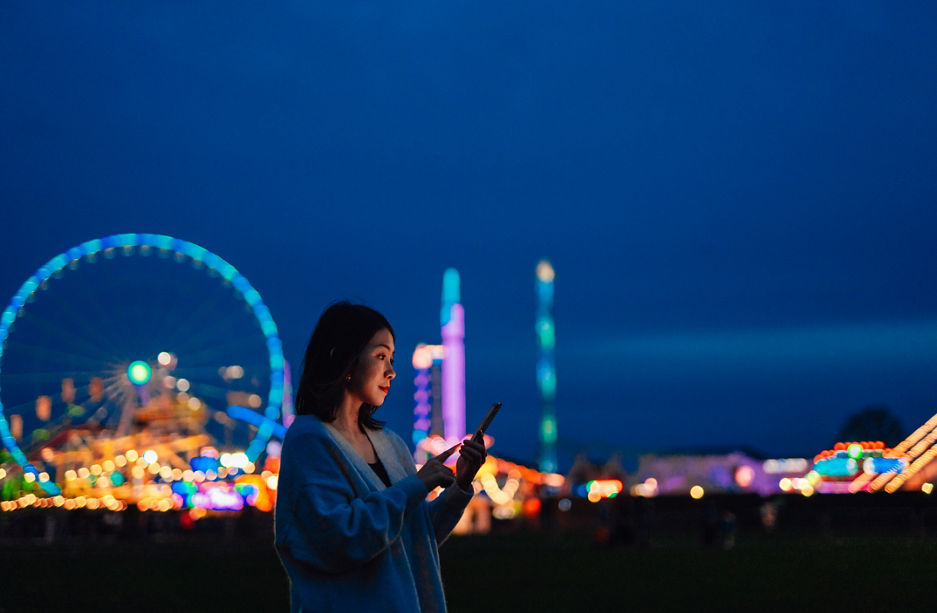 Side view of beautiful young Asian woman looking aways while using smartphone in carnival at night with illuminated night in the background. Mobile banking. Business on the go.
