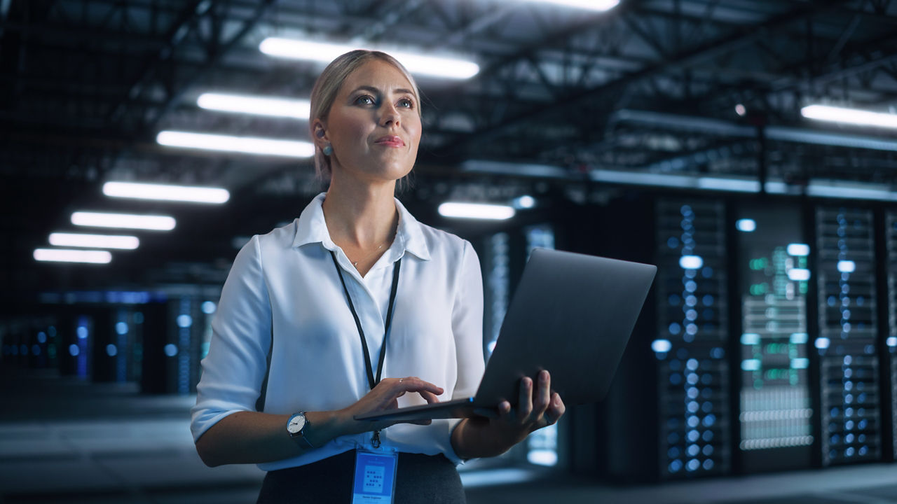 Female Data Center IT Technician Standing at the Server Rack Corridor with Laptop Computer. She is Visually Inspecting Something while Looking at the Screen. Night Office Concept