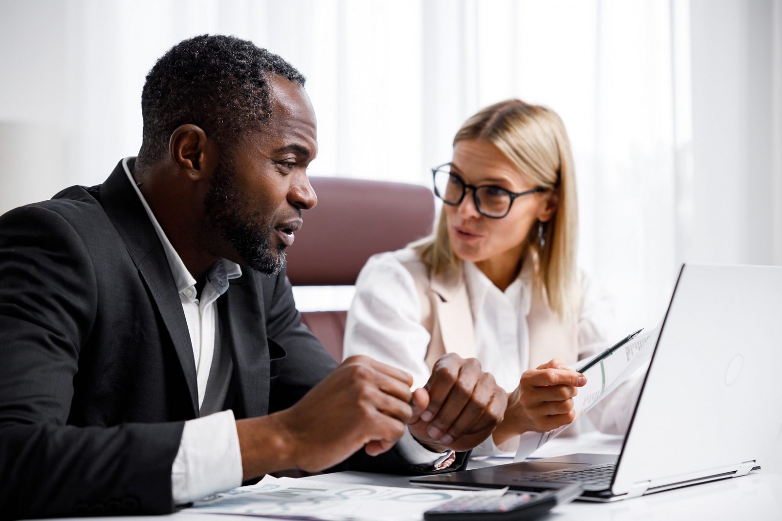 Two people analyze documents on a laptop while sitting at a table in the office. Business partners at work in the office are discussing projects. Multi-racial collaboration