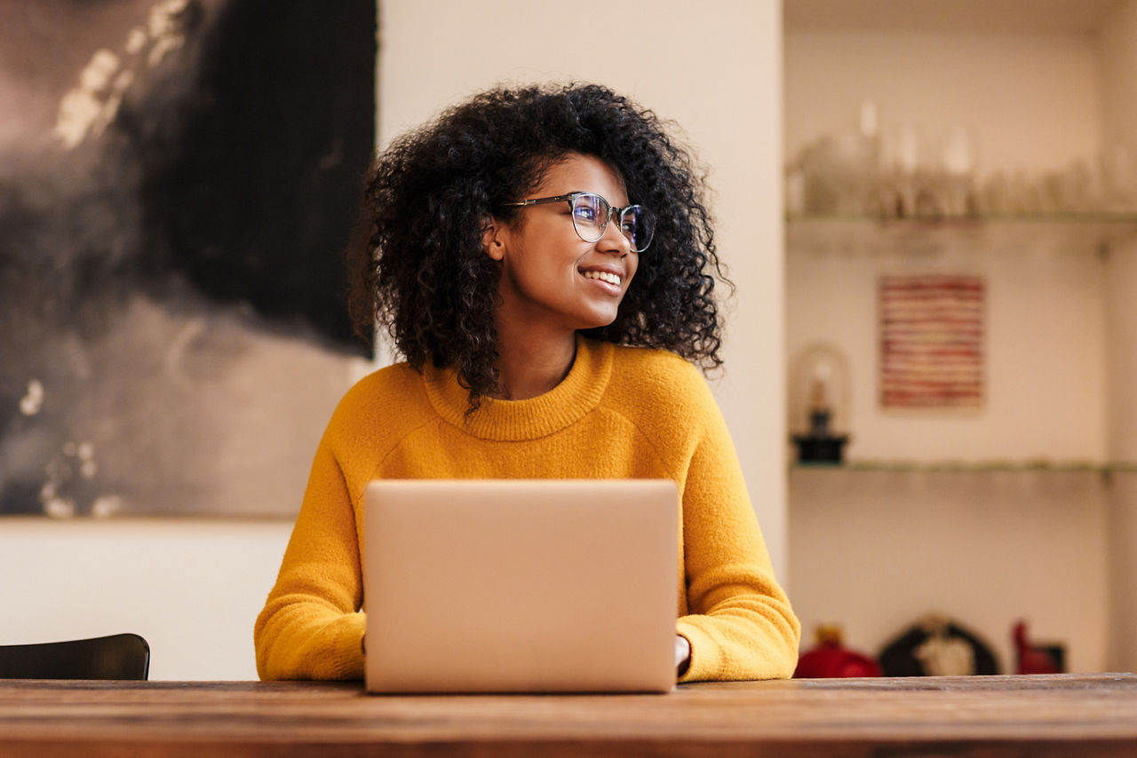 Image of cheerful african american woman in eyeglasses using laptop while sitting at table indoors