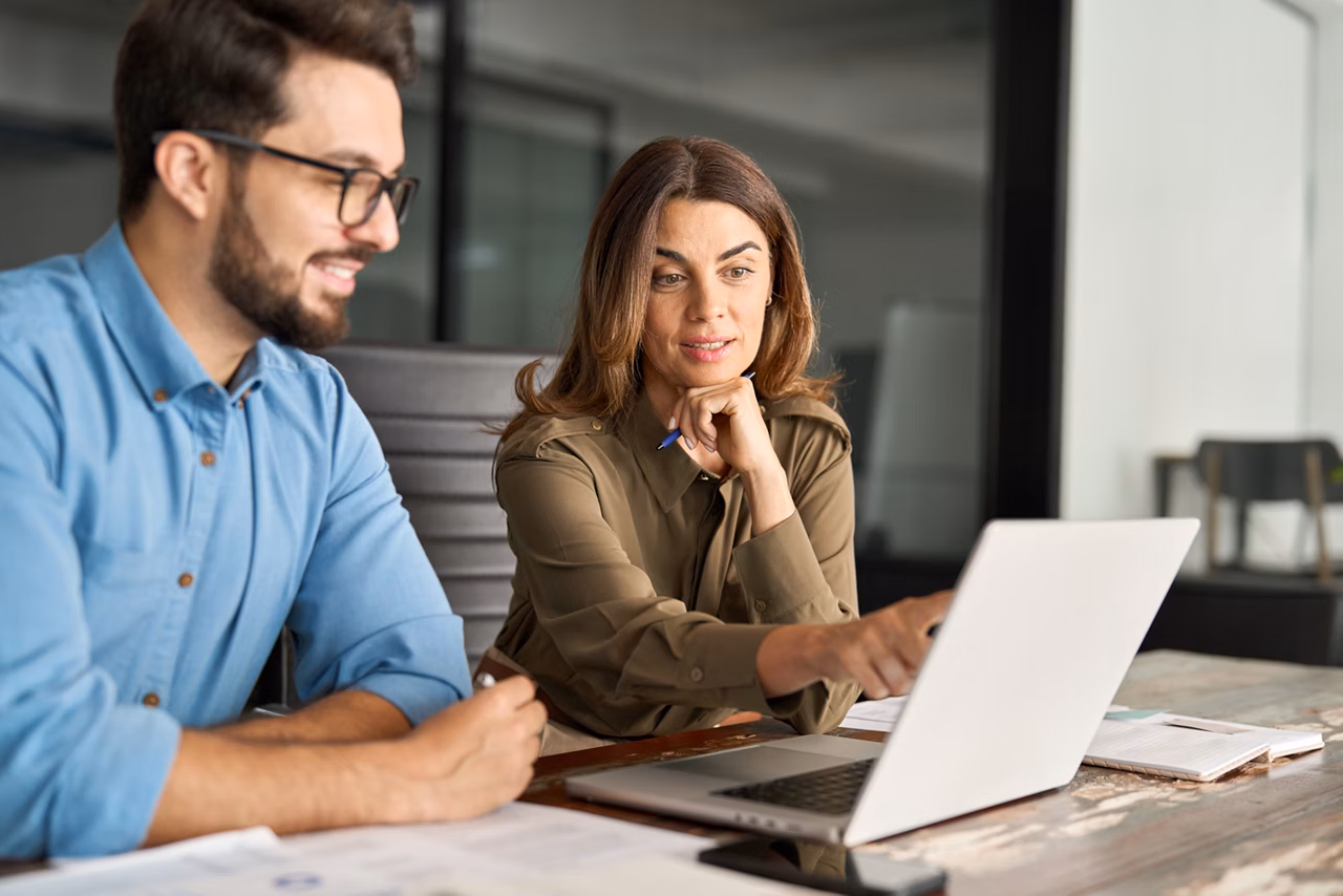 Busy middle aged Latin female manager explaining corporate software work talking to young male employee consulting client working on computer using laptop discussing business plan at office meeting.