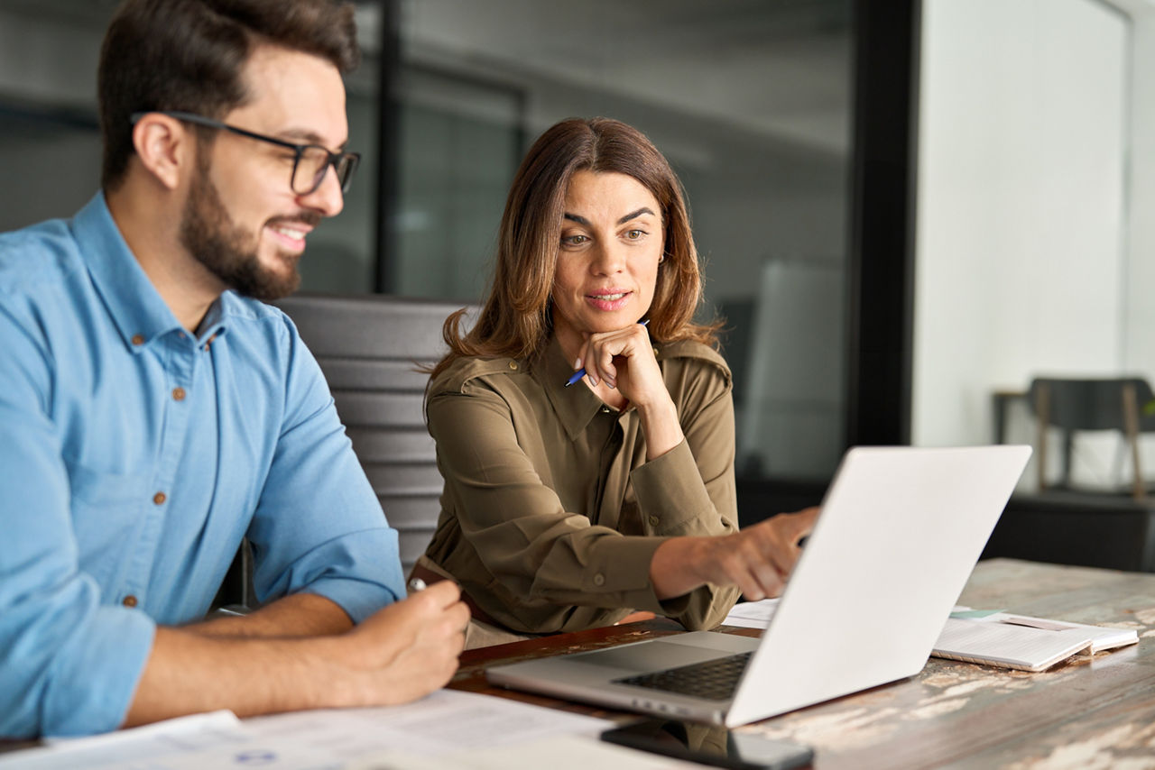 Busy middle aged Latin female manager explaining corporate software work talking to young male employee consulting client working on computer using laptop discussing business plan at office meeting.