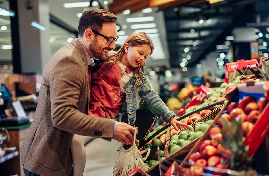 Little girl is buying groceries in the supermarket with her father. He carrying her and picking together fresh fruits