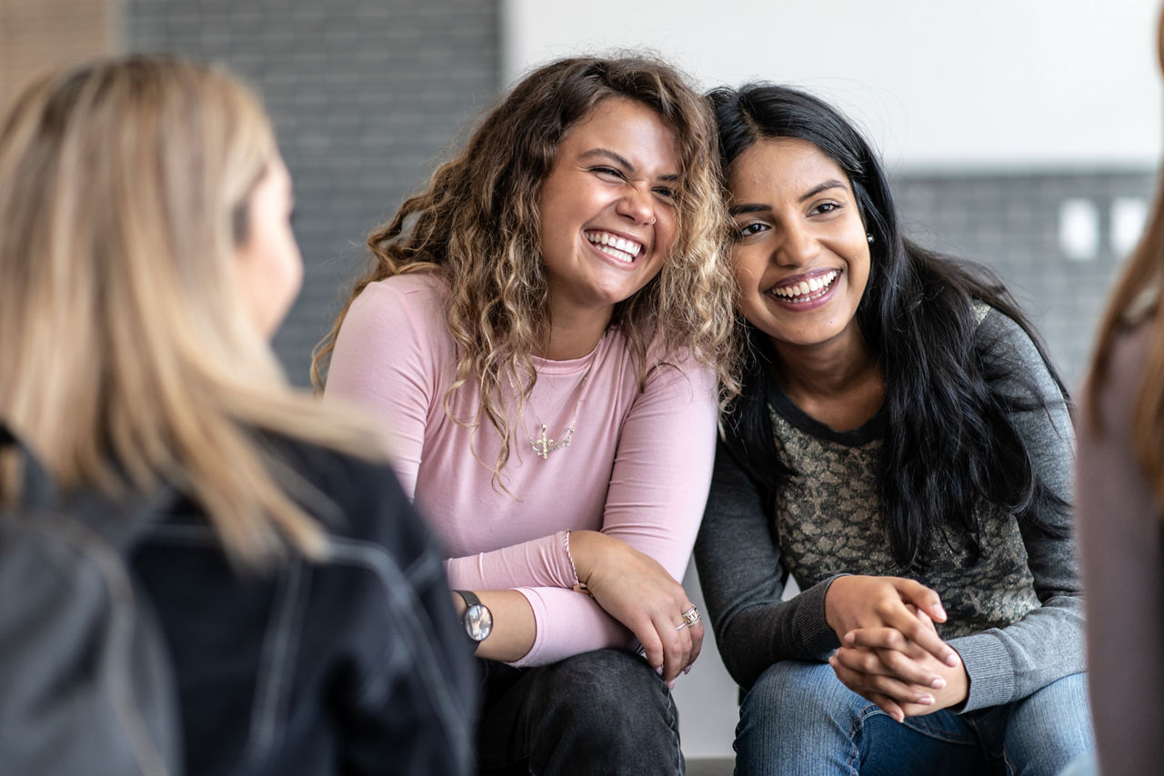 Two female young adults sit closely together, with their heads resting against each others, during a group therapy session.  One is of African decent and the other is of Indian decent.  Bother are dressed casually and are sitting among their multi-ethnic peers with smiles on their faces.