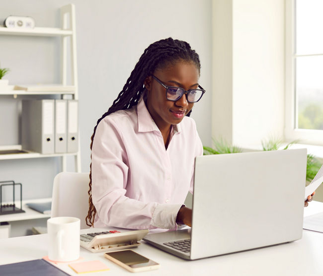 Female accountant at work. Young African American woman in glasses with Afro braids sitting at office desk with cellphone, coffee and calculator working on laptop computer and doing business paperwork