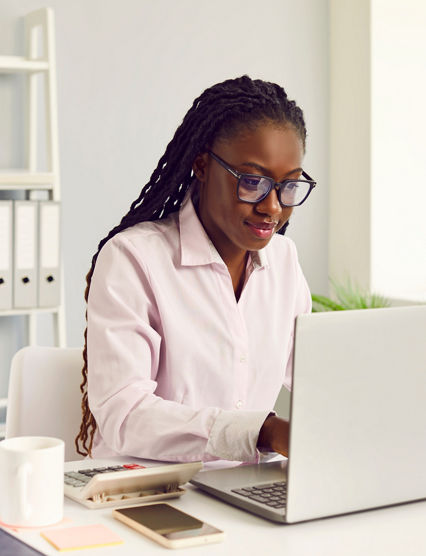 Female accountant at work. Young African American woman in glasses with Afro braids sitting at office desk with cellphone, coffee and calculator working on laptop computer and doing business paperwork