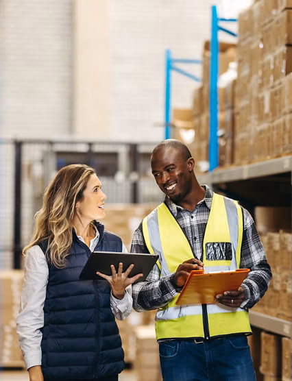 Happy warehouse colleagues smiling at each other while recording inventory. Two cheerful logistics workers using warehouse management software in a modern distribution centre.