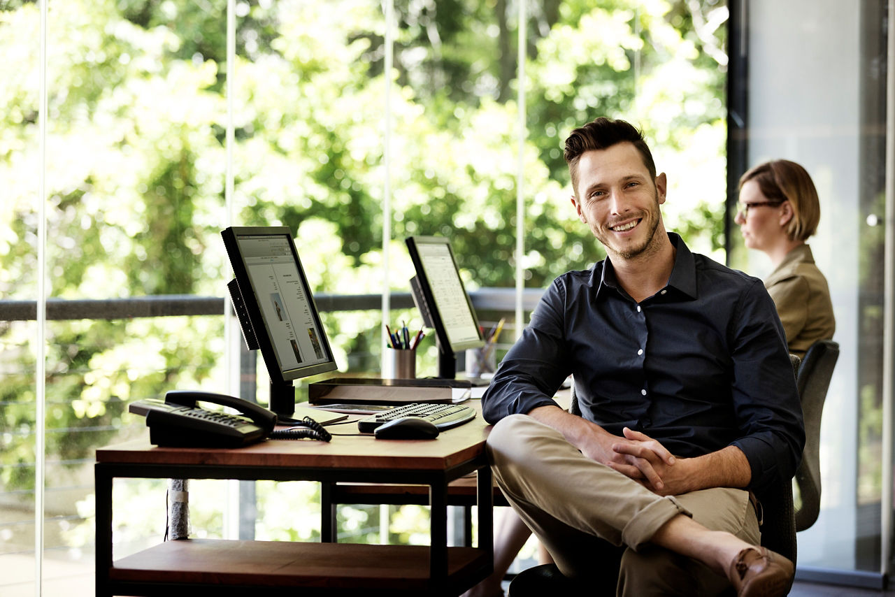 Portrait of happy businessman sitting at computer desk against window in office
