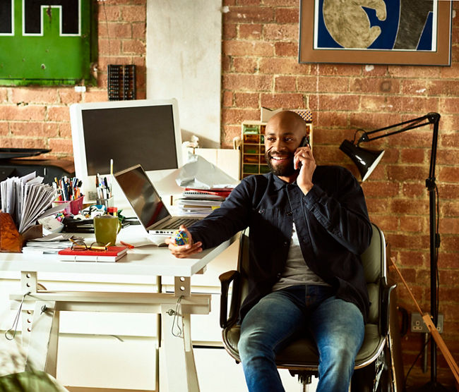 African American small business owner in his 30s talking on smartphone, holding elastic band ball and looking away, confident creative professional in modern studio