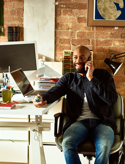 African American small business owner in his 30s talking on smartphone, holding elastic band ball and looking away, confident creative professional in modern studio