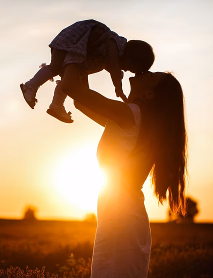 A mother lifts a toddler child in the air above a picturesque sunset sky. A woman and a little girl in a field of lavender flowers