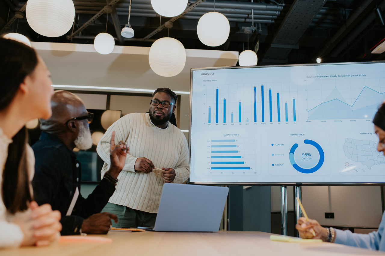A young employee confidently stands in front of a group of colleagues in a modern office environment. He casts a screen with stats and graphs from his laptop onto a large monitor. His colleagues listen intently.