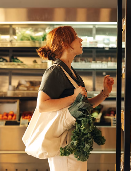 Female shopper choosing food products from a shelf while carrying a bag with vegetables in a grocery store. Young woman doing some grocery shopping in a trendy supermarket.