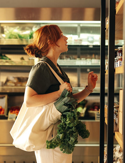 Female shopper choosing food products from a shelf while carrying a bag with vegetables in a grocery store. Young woman doing some grocery shopping in a trendy supermarket.