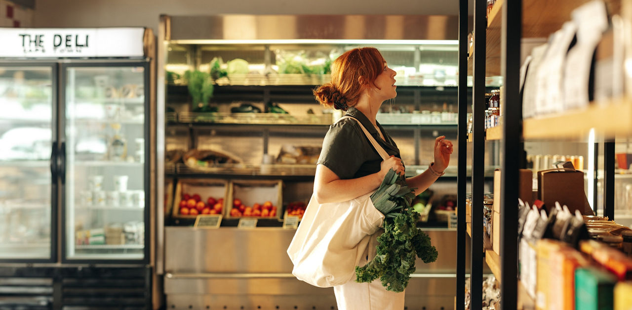 Female shopper choosing food products from a shelf while carrying a bag with vegetables in a grocery store. Young woman doing some grocery shopping in a trendy supermarket.