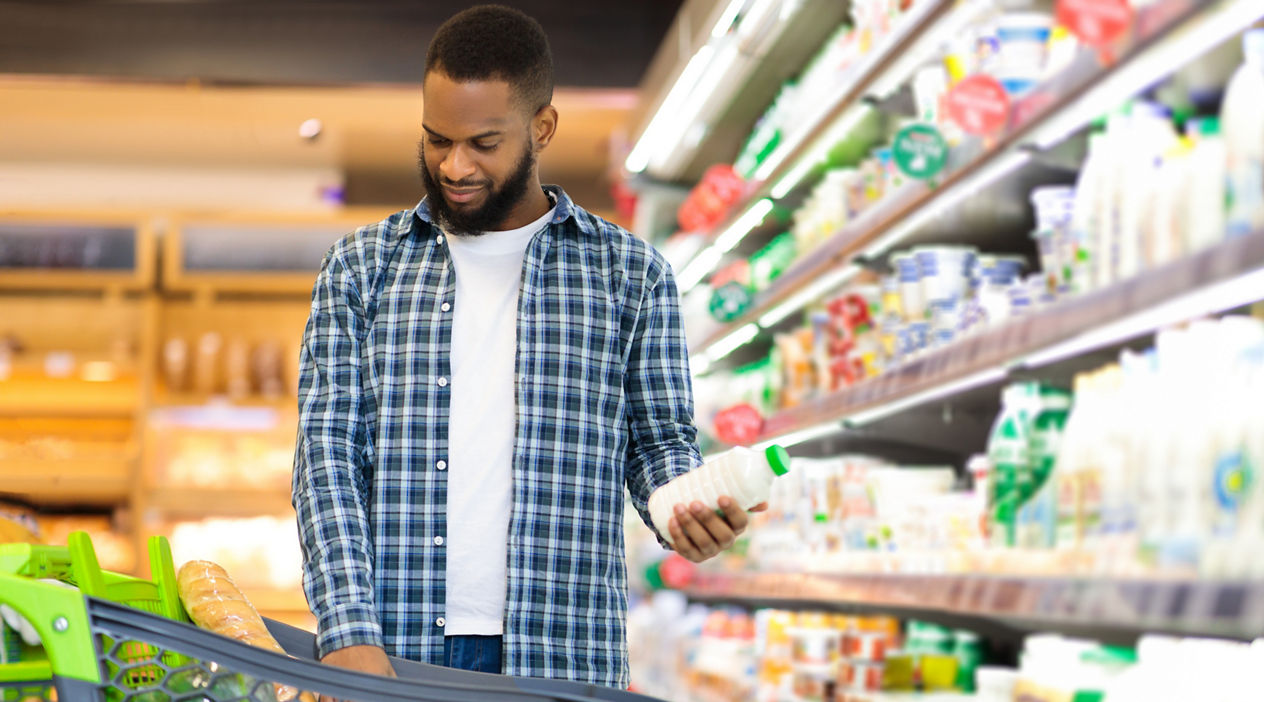 African Man On Grocery Shopping In Supermarket Choosing Dairy Products Buying Food, Standing With Shop Cart Full Of Groceries In Store. Customer In Hypermarket, Coupon And Discount Ad. Free Space