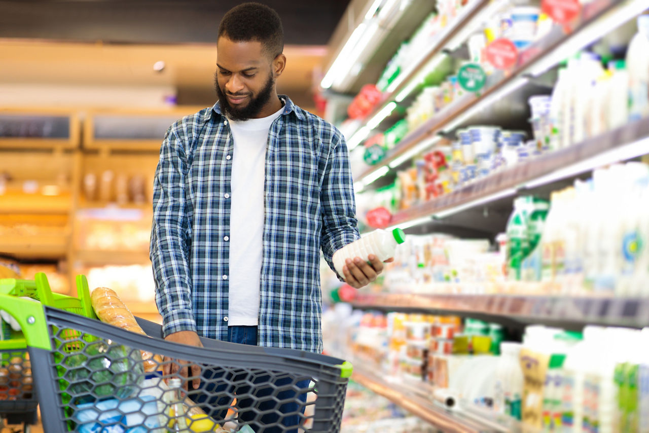 African Man On Grocery Shopping In Supermarket Choosing Dairy Products Buying Food, Standing With Shop Cart Full Of Groceries In Store. Customer In Hypermarket, Coupon And Discount Ad. Free Space