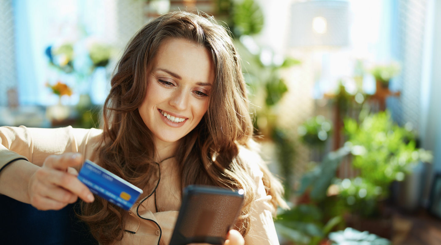 Green Home. happy trendy middle aged woman with long wavy hair with credit card making online shopping on e-commerce website in the modern house in sunny day.